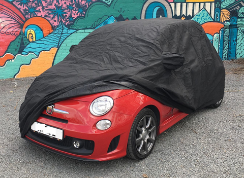 A black indoor car cover fitted on a red car, parked in front of a wall with colorful graffiti.