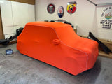 An orange custom fit fleece car cover on a vehicle, displayed in an indoor setting with various car-related decorations in the background.