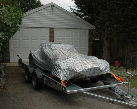 A car cover with a trailer tie down over net is secured on a trailer, parked outdoors.